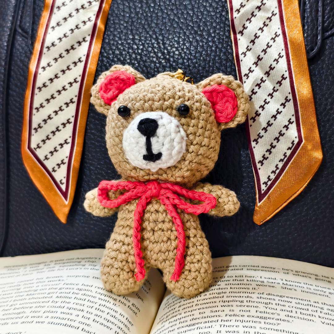 beige teddy on top of a book and beside a orange ribbon on either side in front of a black purse