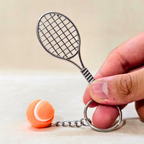 A miniature metal tennis racket keychain with a small orange and white tennis ball charm, held by a hand against a neutral beige background.