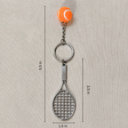A tennis-themed keychain featuring a silver metal tennis racket and key ring, attached to an orange tennis ball charm, shown on a textured beige background with dimension lines and text indicating measurements.