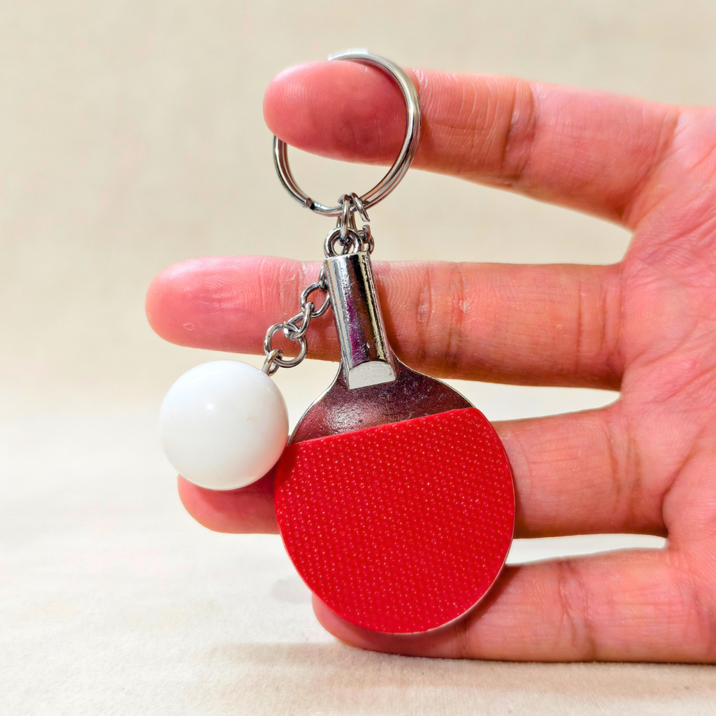 A miniature table tennis-themed keychain held in a hand. The keychain features a small red paddle with a textured surface, a white ball charm, and a silver metal key ring.