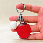 A miniature table tennis-themed keychain held in a hand. The keychain features a small red paddle with a textured surface, a white ball charm, and a silver metal key ring.
