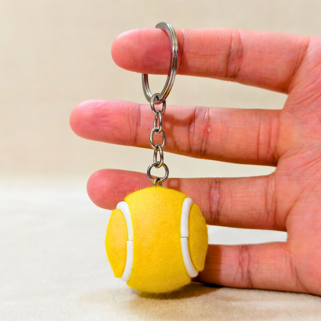 A yellow tennis ball keychain with white lines and a silver metal key ring, held in a person's hand against a neutral beige background.