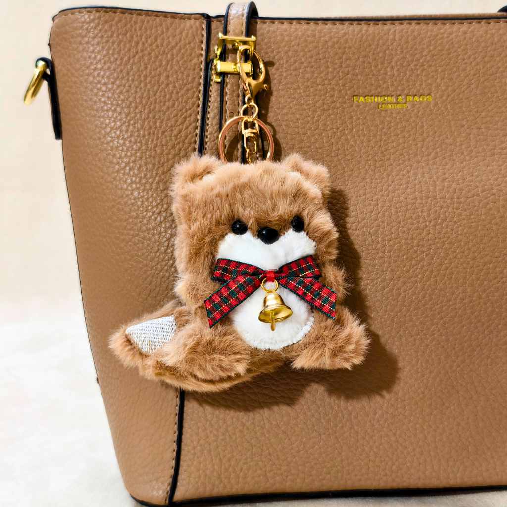 A brown plush teddy bear keychain charm with a red plaid bow and small gold bell, hanging from a gold clasp on a tan textured handbag.