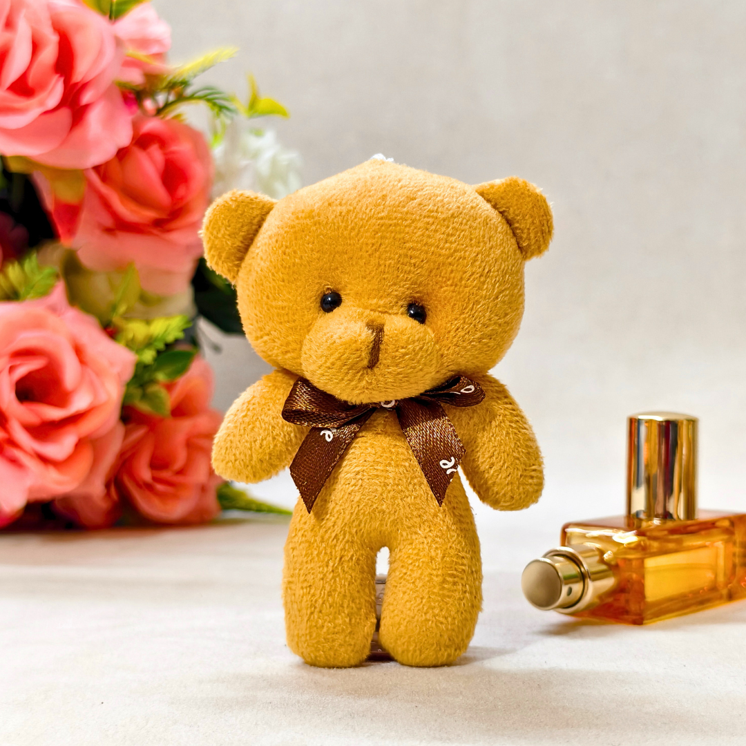 Yellow teddy bear with a brown bow tie next to a perfume bottle and pink flowers on a light background