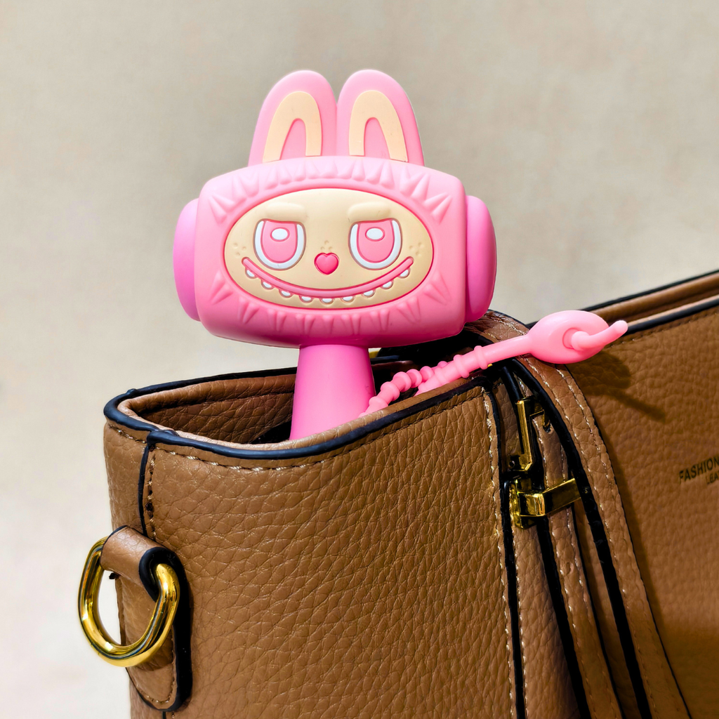 A pink cartoon bunny-shaped accessory with long ears and a coiled pink cord, shown resting inside a brown textured handbag.