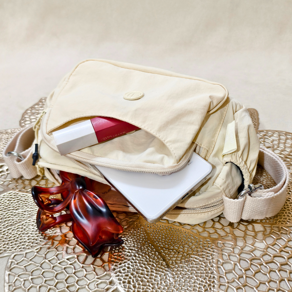 Cream Ivory Sling Bag With Items In Front Of Beige Background.