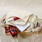 Cream Ivory Sling Bag With Items In Front Of Beige Background.