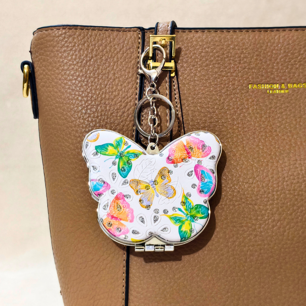 A butterfly-shaped clutch or compact-style bag charm with a white background and colorful butterfly print, attached to a gold-tone key ring and clasp, shown hanging from a brown textured leather bag.