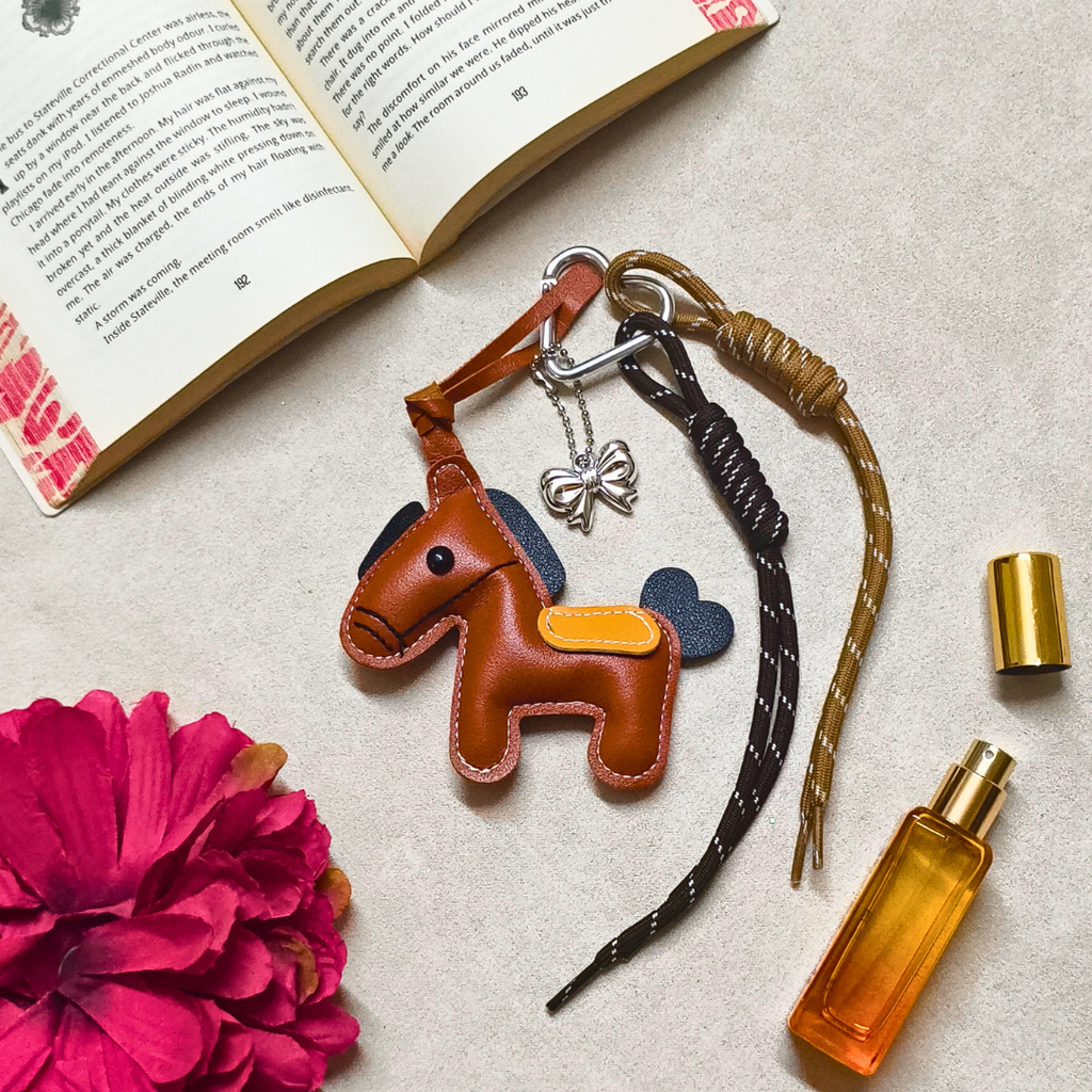 Brown horse-shaped keychain with a bow on a textured surface with a book and pink flower.
