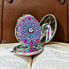 An open silver-toned compact mirror with a colorful mandala design and evil eye motif on the lid, resting on open book pages against a brown leather surface.