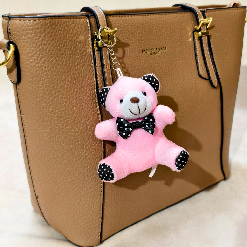 A tan pebbled leather tote bag with gold hardware and a pink plush teddy bear keychain charm featuring black polka-dot ears, bow tie, and paw pads.