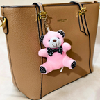 A tan pebbled leather tote bag with gold hardware and a pink plush teddy bear keychain charm featuring black polka-dot ears, bow tie, and paw pads.