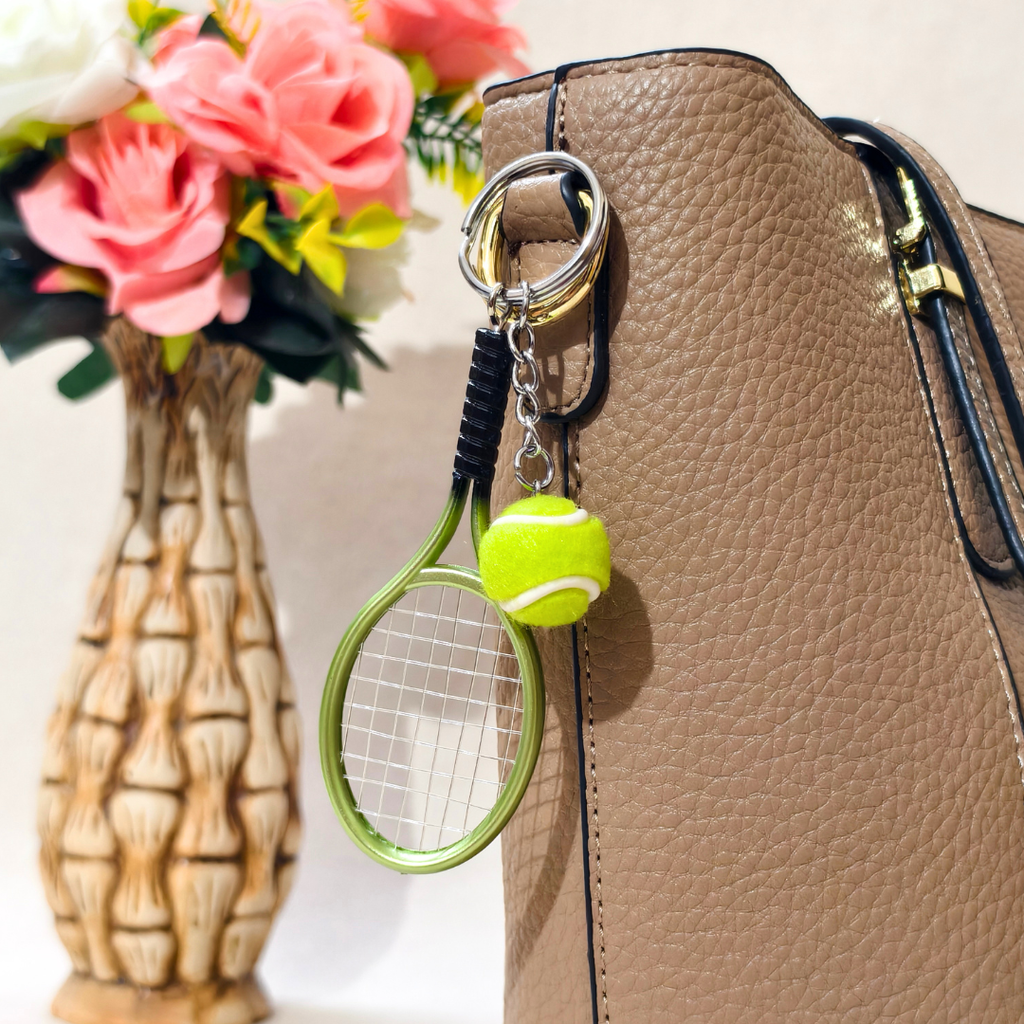 A tennis racket and tennis ball keychain attached to a tan textured handbag, with a vase of pink flowers blurred in the background.