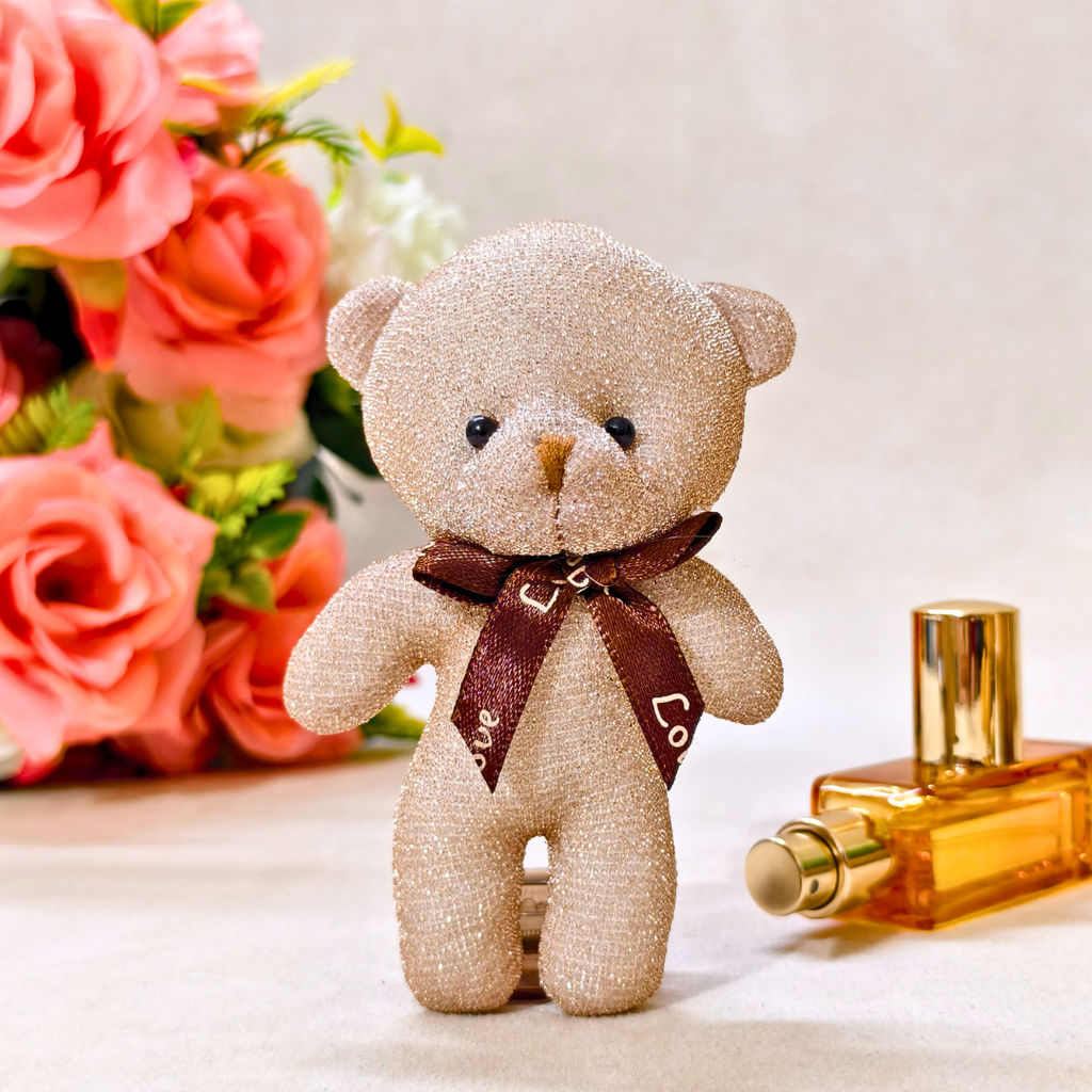 Glittery teddy bear with a brown bow tie next to a perfume bottle and flowers on a light background