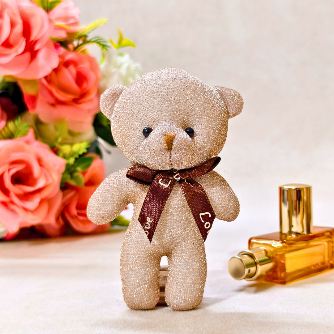 Glittery teddy bear with a brown bow tie next to a perfume bottle and flowers on a light background