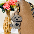A dog-shaped keychain covered in clear rhinestones with a black bow tie and black heart on the chest, hanging from a gold-tone key ring next to a tan textured handbag, with a vase of pink flowers blurred in the background.