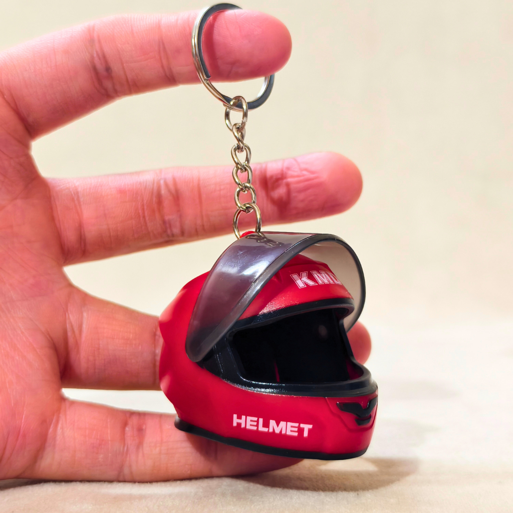 A miniature red motorcycle helmet keychain held in a person's hand. The keychain features a small red helmet with a clear visor, black trim, and a silver metal key ring and chain.
