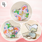 A butterfly-shaped compact mirror with a silver-tone metal frame and keychain ring, shown closed and open in a collage on a soft pink background. The butterfly surface is white with colorful butterfly prints in pink, orange, yellow, green, and blue, decorated with small rhinestones.