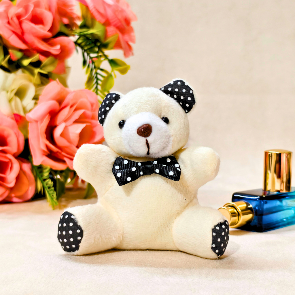 A small plush teddy bear perfume bottle with a cream body, black and white polka dot ears, nose, and bow tie, sitting on a beige surface with pink roses and a perfume bottle in the background.