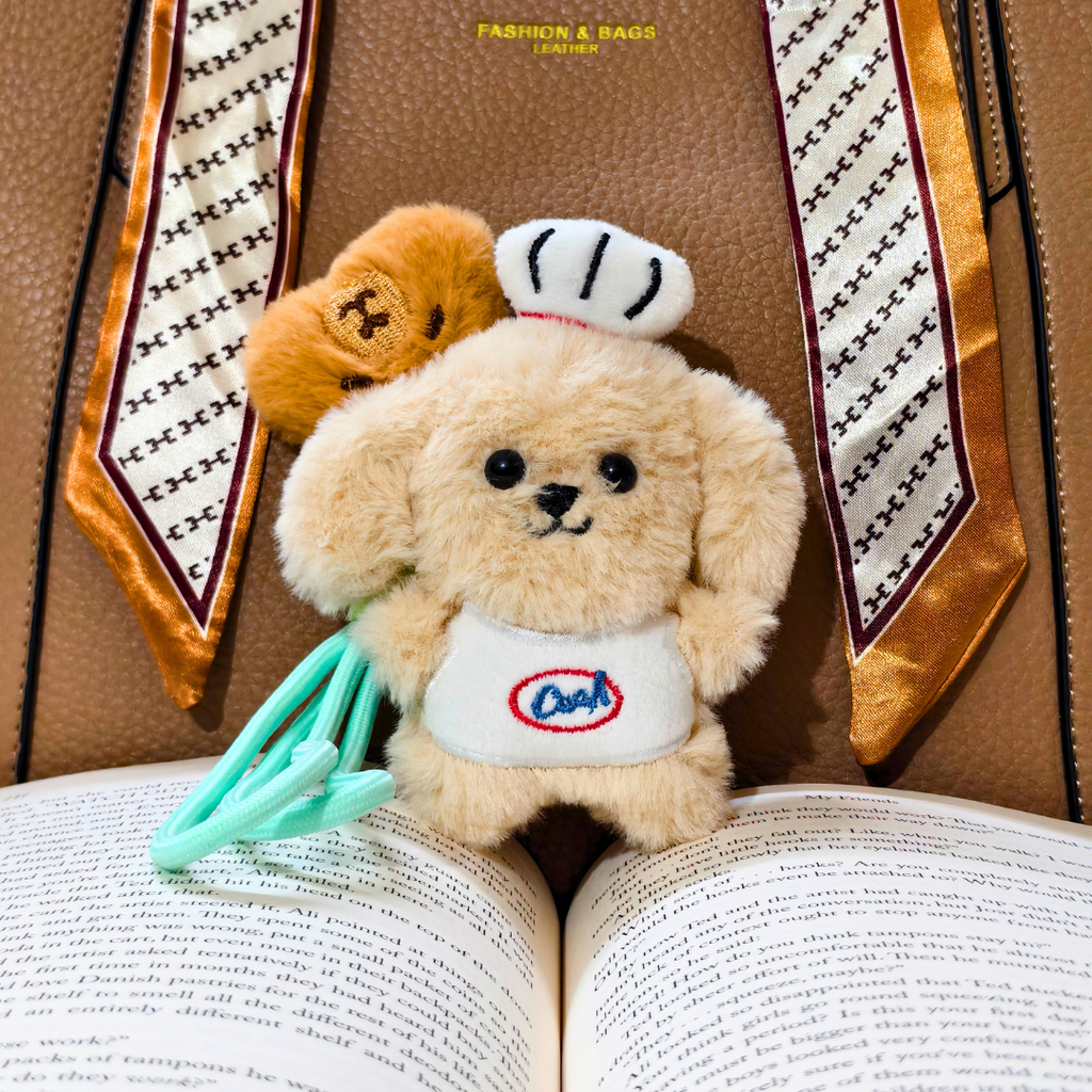 A small plush dog keychain or charm styled on an open book, wearing a white chef hat and bib with a blue and red logo, holding mint green chopsticks. A second brown plush dog is tucked behind it.
