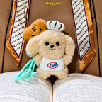 A small plush dog keychain or charm styled on an open book, wearing a white chef hat and bib with a blue and red logo, holding mint green chopsticks. A second brown plush dog is tucked behind it.