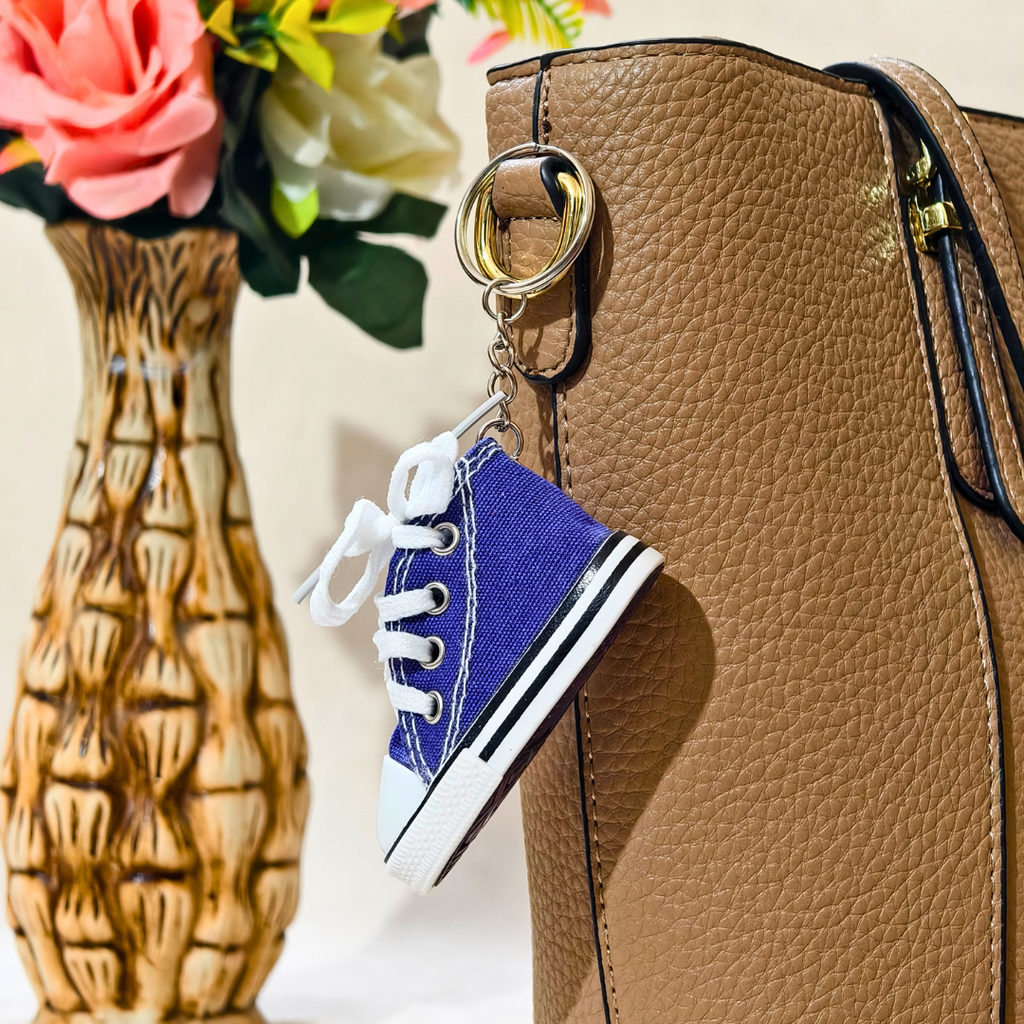 A miniature blue canvas sneaker keychain charm with white laces and a black-and-white sole, hanging from a gold ring on a brown textured handbag. A decorative vase with pink and white flowers is blurred in the background.