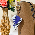 A miniature blue canvas sneaker keychain charm with white laces and a black-and-white sole, hanging from a gold ring on a brown textured handbag. A decorative vase with pink and white flowers is blurred in the background.