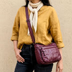 Person wearing a mustard yellow shirt, dark jeans, and a white scarf, holding a purple leather bag against a beige wall.