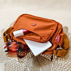 Rust Brown Sling Bag With Items In Front Of Beige Background.