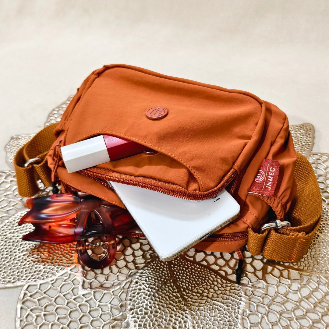 Rust Brown Sling Bag With Items In Front Of Beige Background.