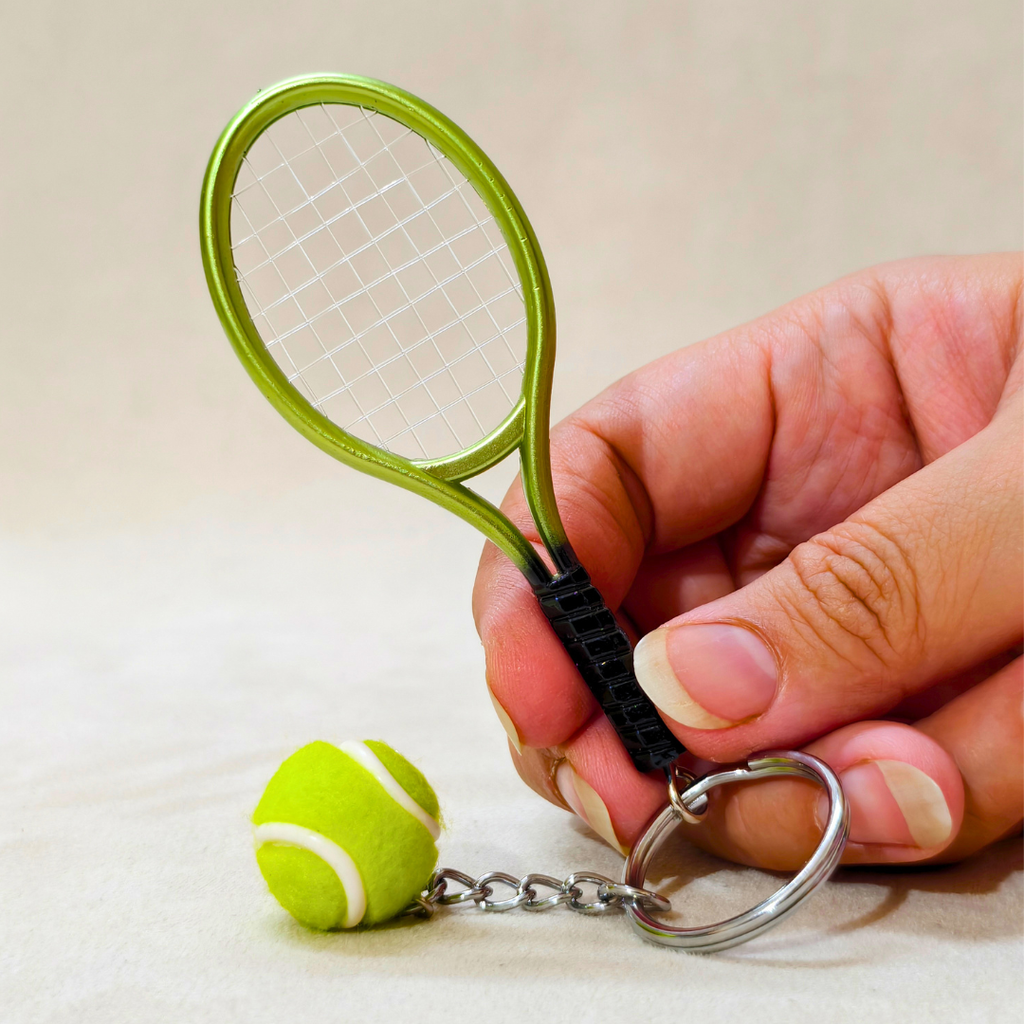 A miniature tennis racket keychain with a small tennis ball charm, held by a hand against a neutral beige background.