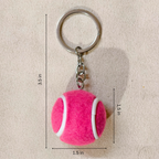 A pink felt tennis ball keychain with a silver metal split ring and chain, shown on a beige fabric background with dimension lines and text indicating measurements of 3.5 in height, 1.