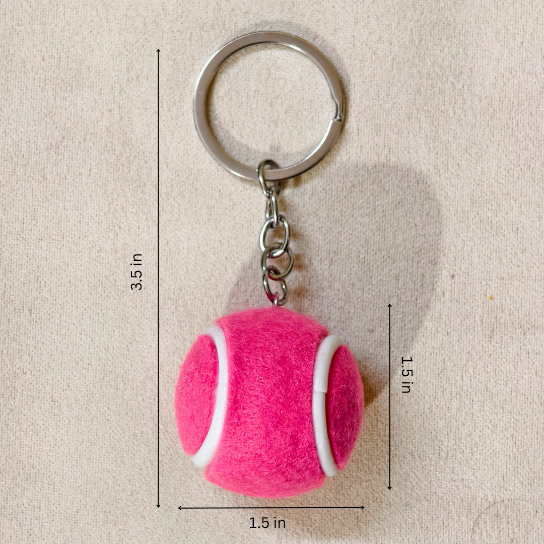 A pink felt tennis ball keychain with a silver metal split ring and chain, shown on a beige fabric background with dimension lines and text indicating measurements of 3.5 in height, 1.