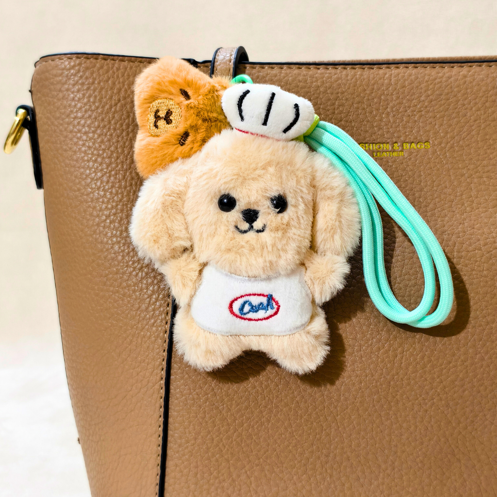 A plush dog-shaped keychain or bag charm with a mint green wrist strap, attached to a brown textured handbag. The charm features a cream-colored dog wearing a white apron with 'Dad' embroidered in red and blue, with a small orange-brown bear plush tucked behind it.