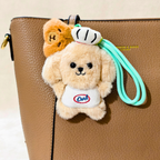A plush dog-shaped keychain or bag charm with a mint green wrist strap, attached to a brown textured handbag. The charm features a cream-colored dog wearing a white apron with 'Dad' embroidered in red and blue, with a small orange-brown bear plush tucked behind it.