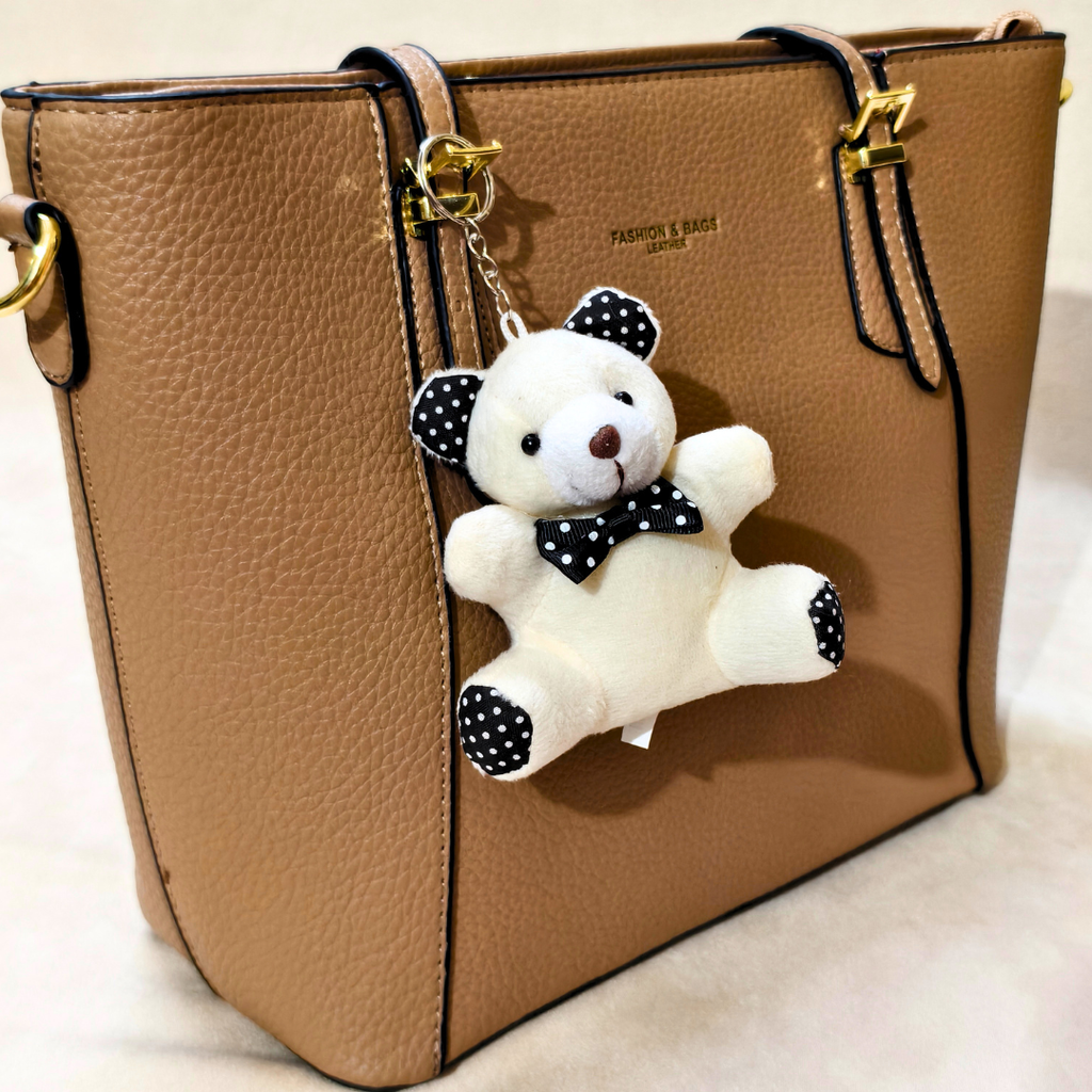 A tan pebbled leather handbag with gold-tone hardware and a hanging plush keychain. The keychain is a white teddy bear with black polka-dot ears, bow tie, and paw pads, attached with a gold ring.