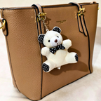 A tan pebbled leather handbag with gold-tone hardware and a hanging plush keychain. The keychain is a white teddy bear with black polka-dot ears, bow tie, and paw pads, attached with a gold ring.