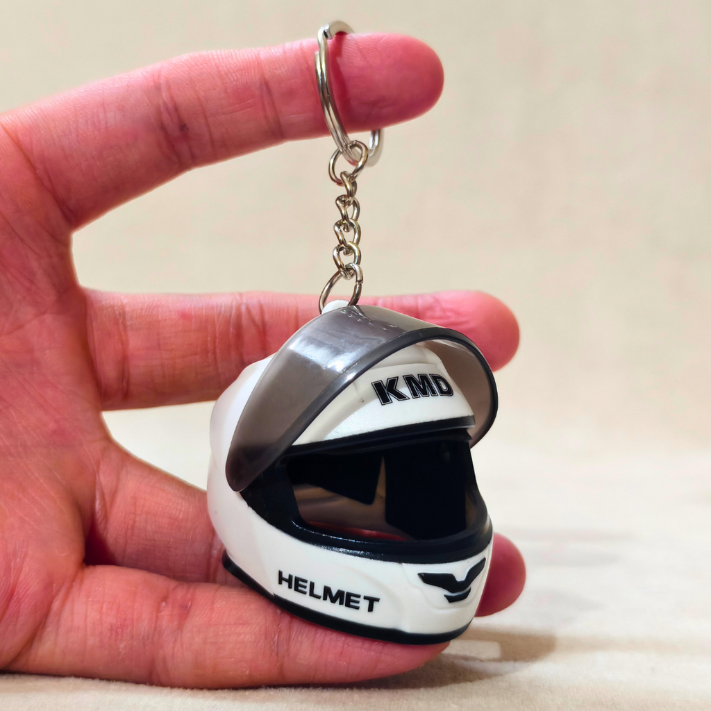 A miniature white and black motorcycle helmet keychain held in a person's hand. The keychain features 'KMD' and 'HELMET' printed on it, attached to a silver metal split ring and chain.