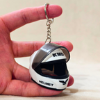 A miniature white and black motorcycle helmet keychain held in a person's hand. The keychain features 'KMD' and 'HELMET' printed on it, attached to a silver metal split ring and chain.