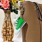 A miniature green canvas sneaker keychain charm with white laces and a black stripe, attached to a gold key ring and shown hanging next to a brown textured handbag.