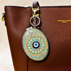 An oval-shaped decorative keychain charm with a colorful mandala pattern and a blue evil eye in the center, hanging from a gold-tone ring on a brown textured leather bag.