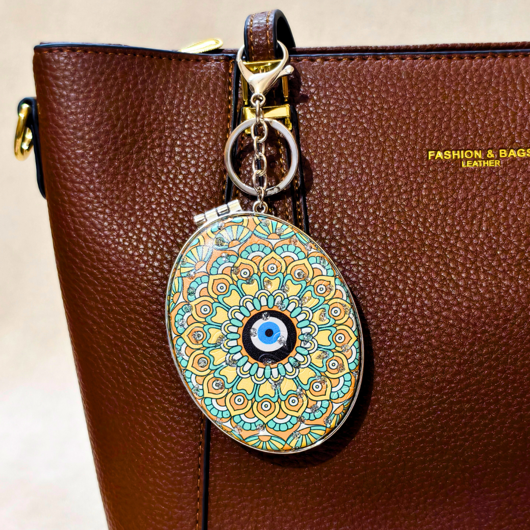 An oval-shaped decorative keychain charm with a colorful mandala pattern and a blue evil eye in the center, hanging from a gold-tone ring on a brown textured leather bag.