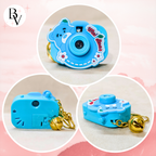 A small blue toy camera keychain shown from three angles in circular frames on a soft pink background. The camera has a cartoon bear face, white and red flower details, decorative script text, and a gold bell attached by a short gold chain.