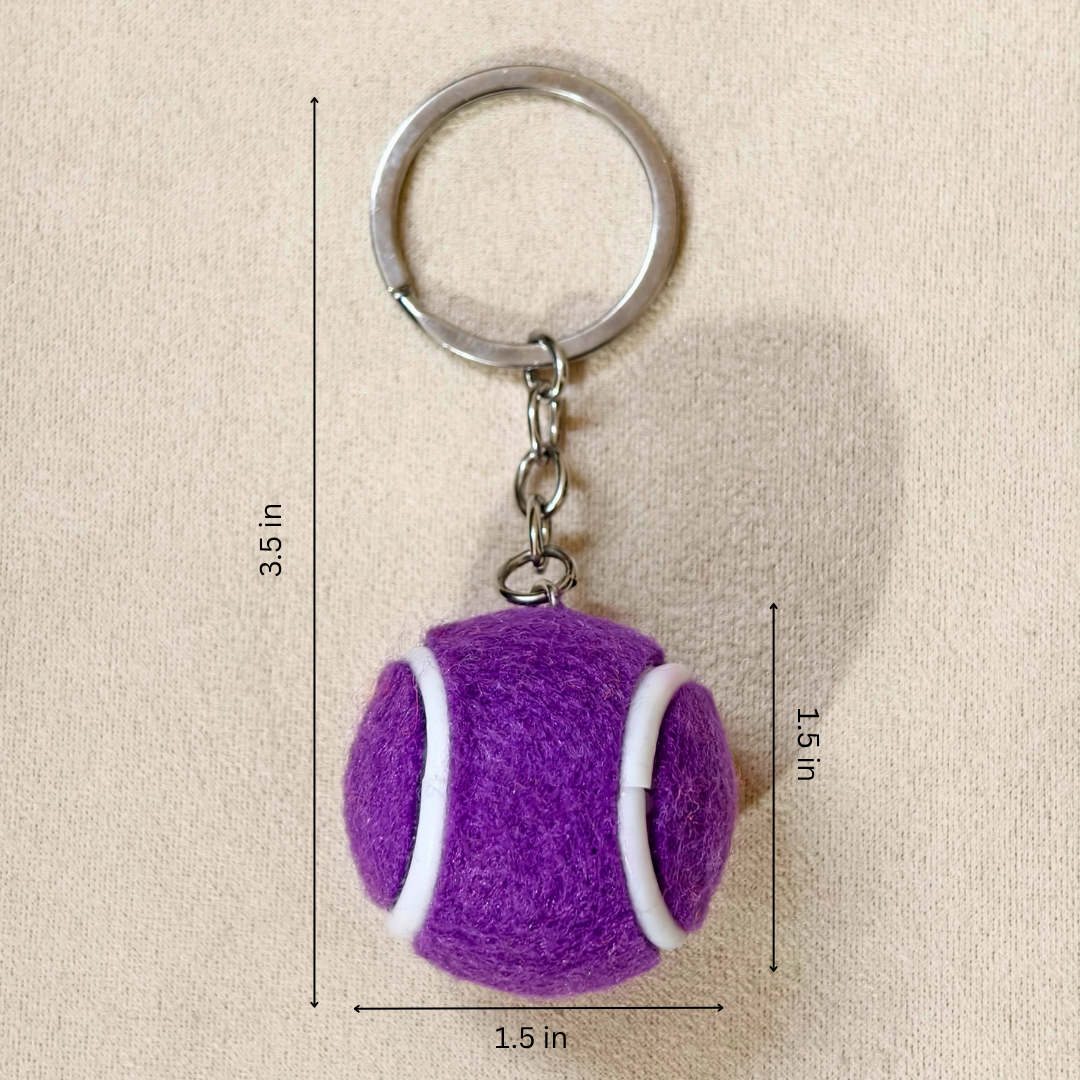 Purple felt tennis ball keychain with white curved accents and a silver metal key ring, shown on a beige fabric background with dimension lines indicating 1.5 in height, 1.