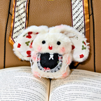 A plush white mouse toy with embroidered red strawberries, pink cheeks, and a black mouth, resting on an open book in front of a patterned box.