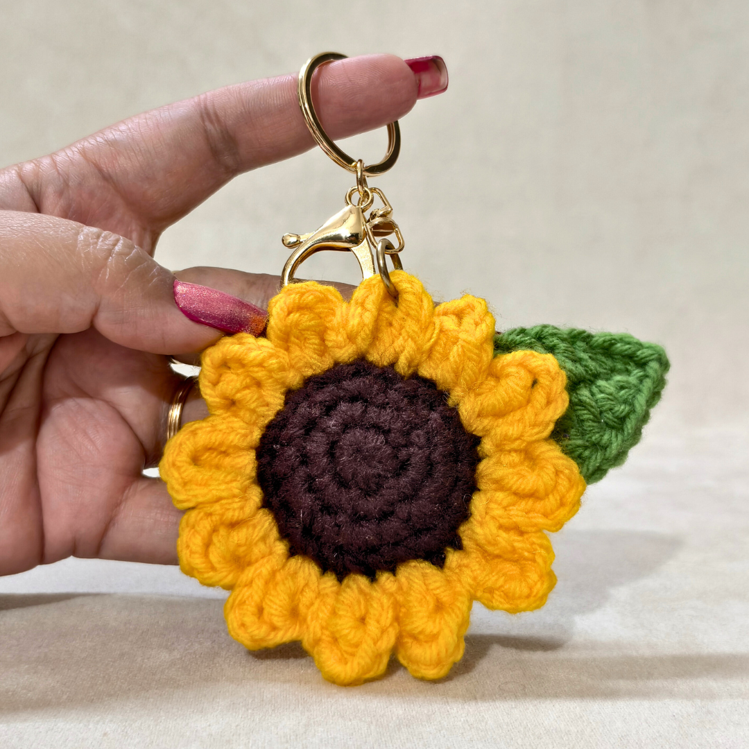 Hand holding a crocheted sunflower keychain with a green leaf on a neutral background