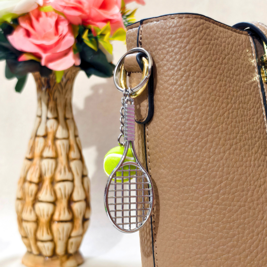 A silver metal tennis racket keychain with a neon yellow tennis ball charm, attached to a black ring on a brown textured leather handbag. In the background, a patterned ceramic vase holds pink artificial roses.