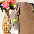 A silver metal tennis racket keychain with a neon yellow tennis ball charm, attached to a black ring on a brown textured leather handbag. In the background, a patterned ceramic vase holds pink artificial roses.