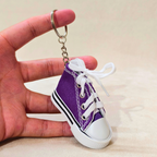 A miniature purple canvas high-top sneaker keychain with white laces and a white rubber toe cap, held by a hand against a light beige background.