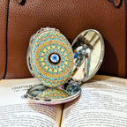 An oval compact mirror with a colorful mandala pattern and a central blue evil eye, shown open on top of open book pages against a brown leather background.
